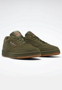 Un paio di sneakers Reebok verde oliva con suole in gomma, toppe con logo Reebok e lacci su uno sfondo bianco.