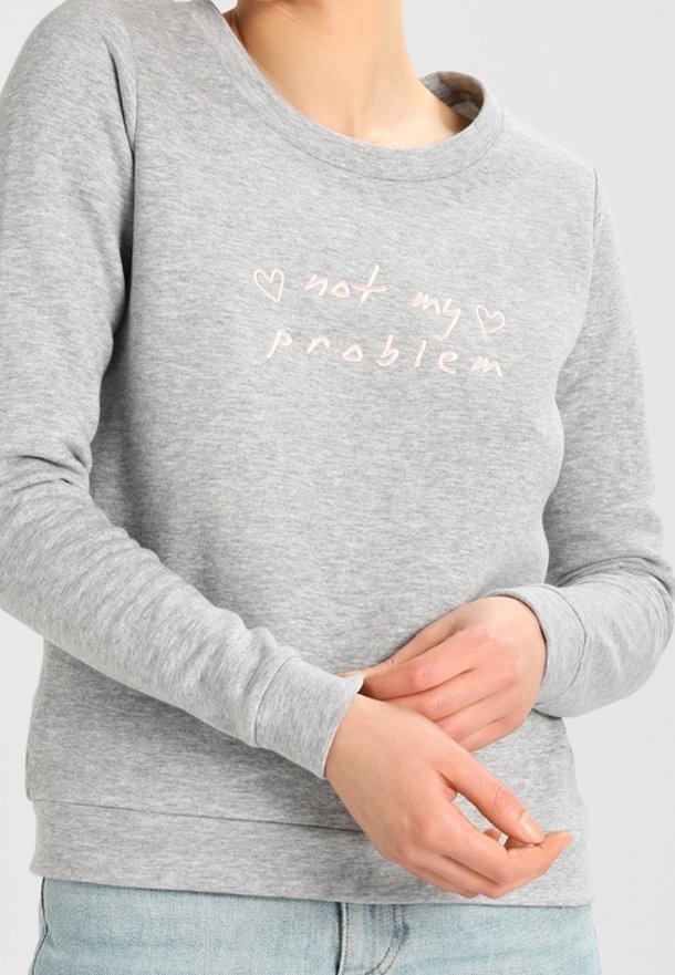 Personne portant un sweat-shirt gris avec l'inscription « ce n'est pas mon problème » et deux contours de cœur brodés en rose clair, les mains jointes à la taille.