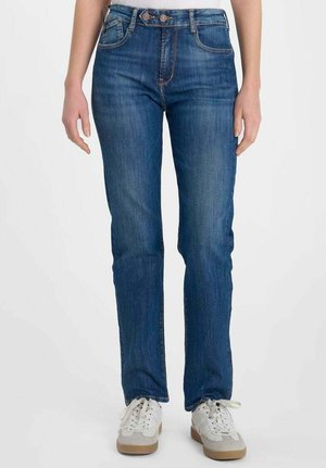 Femme portant un jean droit bleu et des baskets blanches, debout devant un fond uni.