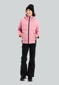 Persona sonriente que lleva una chaqueta acolchada rosa, un gorro negro, pantalones negros con estampado de estrellas y botas negras, de pie con las manos en los bolsillos.