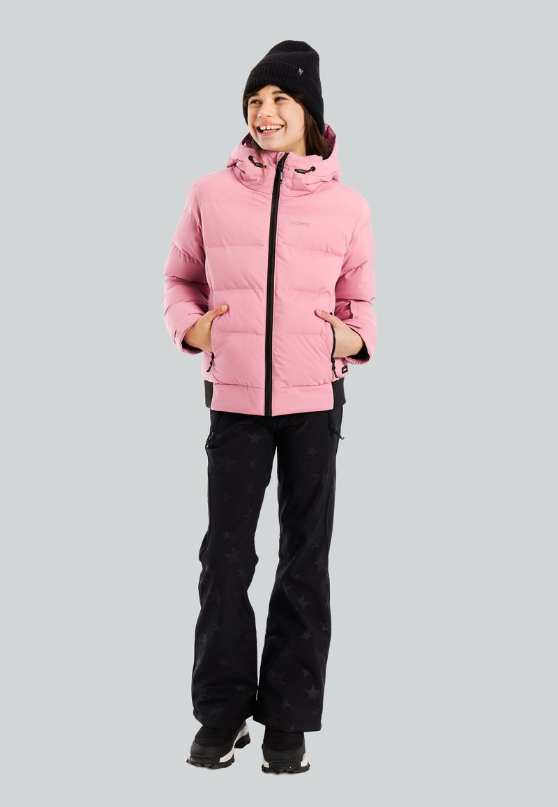 Persona sonriente que lleva una chaqueta acolchada rosa, un gorro negro, pantalones negros con estampado de estrellas y botas negras, de pie con las manos en los bolsillos.