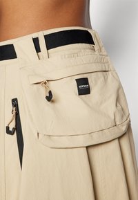 Beige szoknya nadrág puha textúrával, rugalmas fekete derékkal, oldalsó cipzárokkal és zsebkendő zsebekkel, amelyek húzózsinórral záródnak.