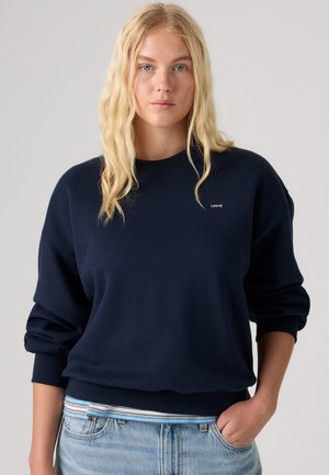 Lyshåret kvinde med bølget hår iført en navyblå Levi's sweatshirt over en stribet skjorte og lyseblå jeans, stående mod en ensfarvet baggrund.