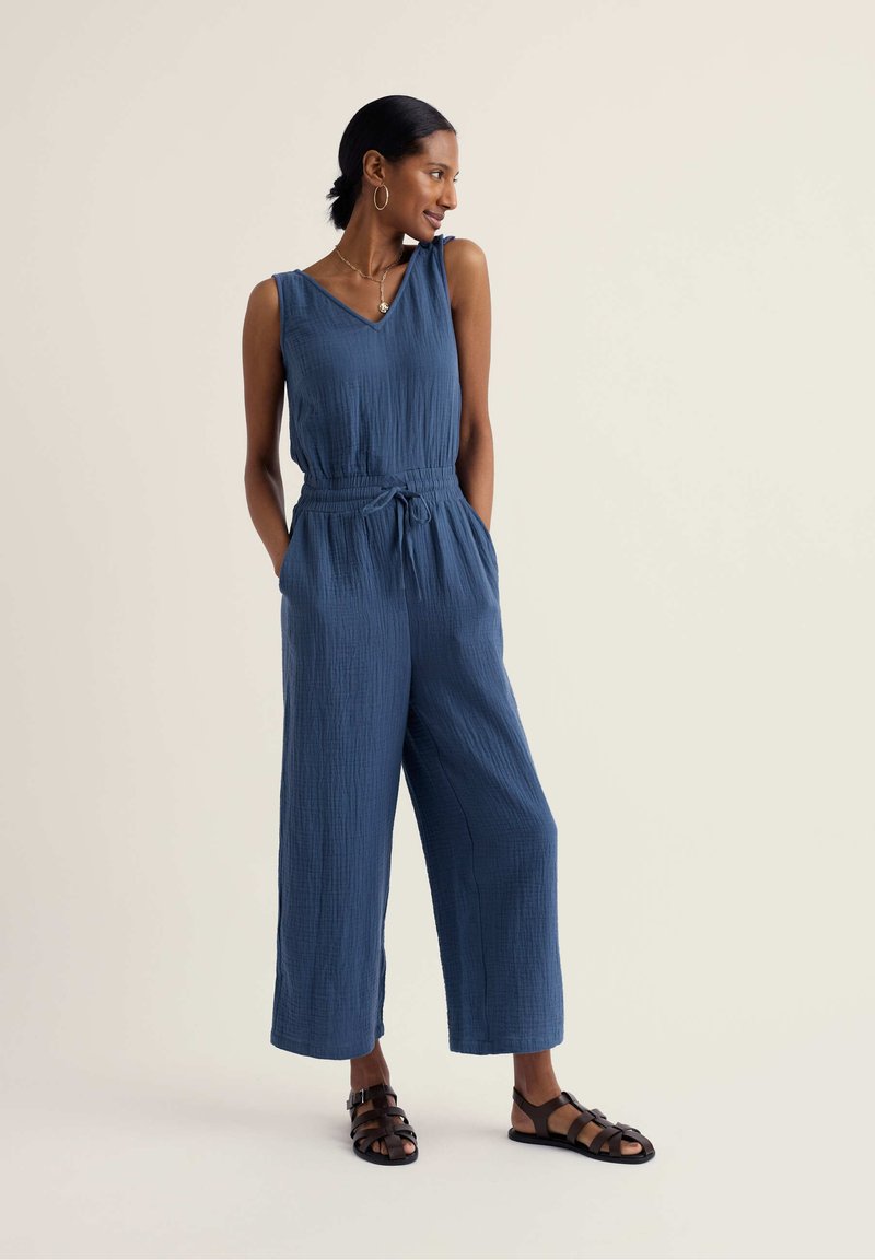 Frau steht, trägt einen blauen ärmellosen Jumpsuit mit Taschen und schwarze Sandalen, schaut seitlich vor einem einfarbigen Hintergrund.