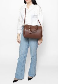 Borsa a mano in pelle marrone con un design strutturato, doppi manici, tracolla rimovibile e un cartellino per i bagagli attaccato.