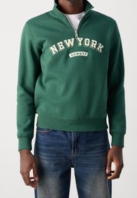 Muž v zelené mikině se zipem do půlky a nápisem "NEW YORK SCHOTT" a modrých džínách, stojící před jednobarevným světlým pozadím.