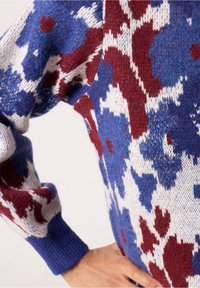 Pletený svetr s modrými, červenými a bílými maskáčovými vzory; žebrované manžety; měkká textura; detailní záběr rukávu s propracovanými detaily.