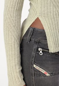 Pull vert clair côtelé à manches longues, associé à un jean gris foncé avec une poche zippée et une étiquette de marque.