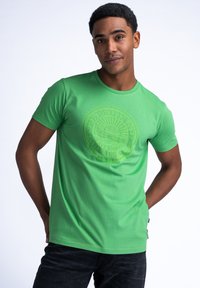 Camiseta de algodón verde con un logo redondo texturizado centrado en el pecho. Mangas cortas y cuello redondo, combinada con jeans oscuros.