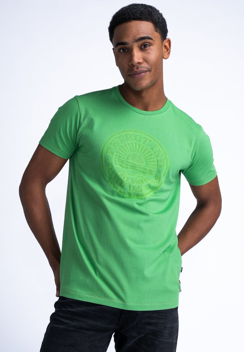 Camiseta de algodón verde con un logo redondo texturizado centrado en el pecho. Mangas cortas y cuello redondo, combinada con jeans oscuros.