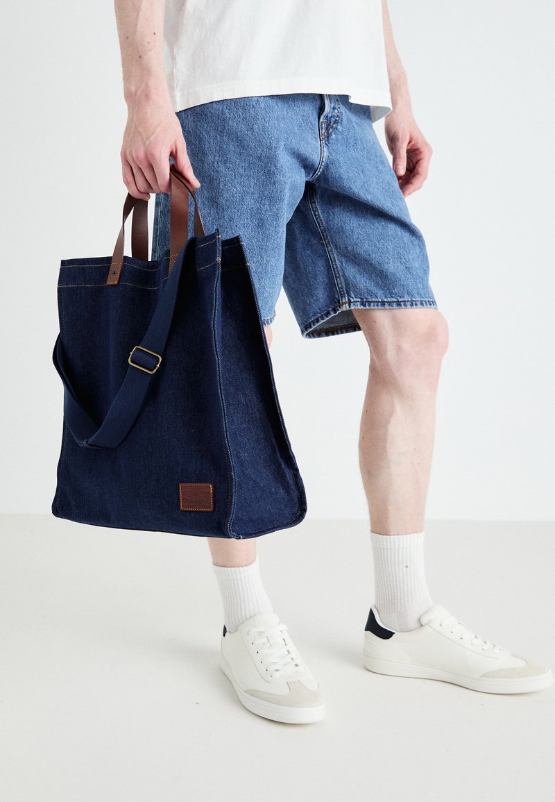Marineblauwe denim draagtas met bruine leren handvatten en een schouderriem, voorzien van contrasterende stiksels en een klein leren label.