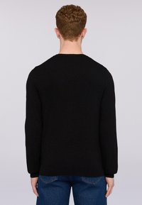 Černý svetr s dlouhým rukávem a kulatým výstřihem, hladká textura a žebrované manžety, v kombinaci s modrými džíny. Pohled zezadu.