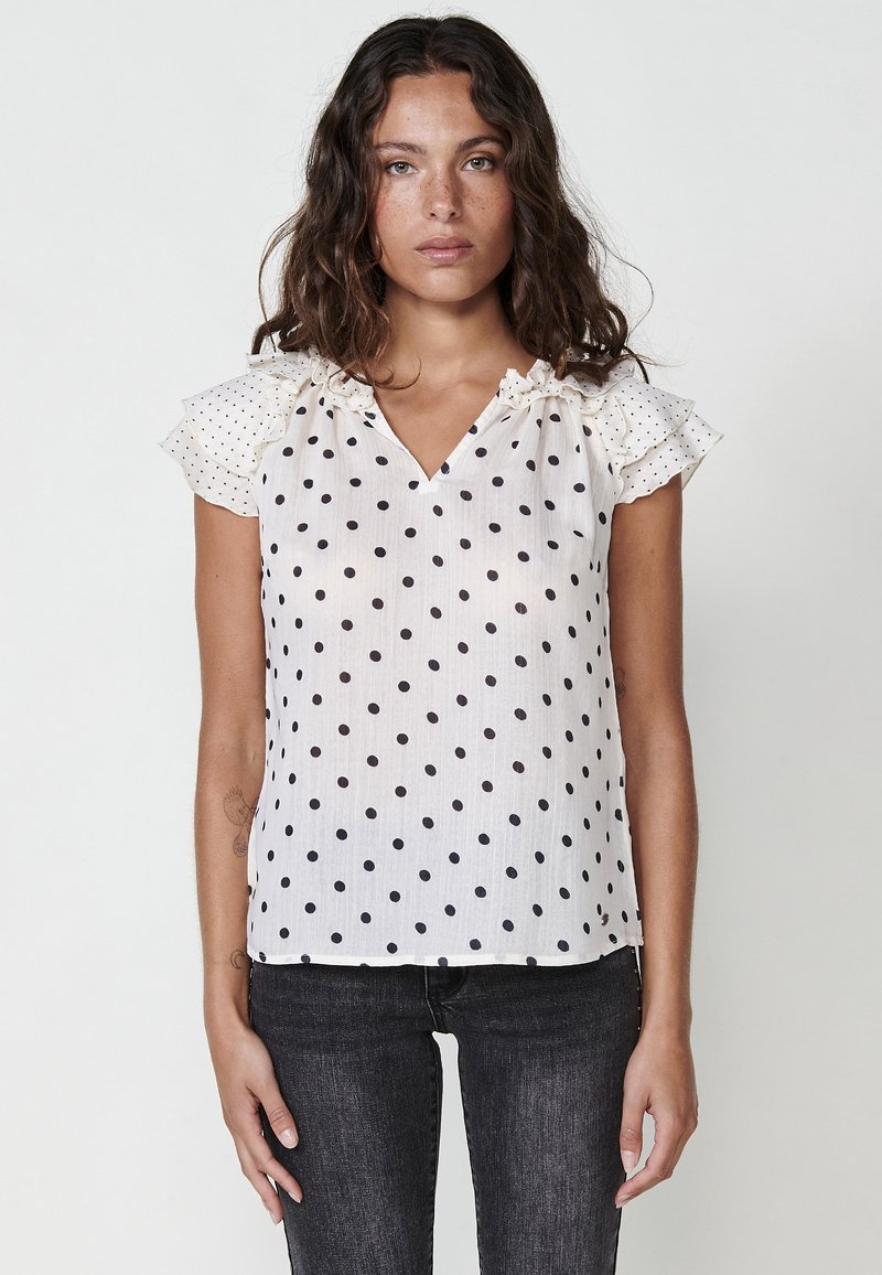 Blusa bianca con pois neri, scollatura a V e maniche corte con volant. Tessuto leggero, vestibilità rilassata e orlo che cade sull'anca.