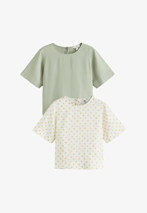 Femme portant des lunettes de soleil marron, un haut à manches courtes vert clair, une jupe blanche longueur genou et un grand bracelet, debout devant un fond blanc uni.