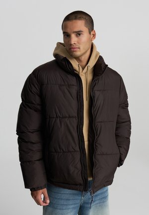 Giacca puffer marrone scuro con chiusura a zip, caratterizzata da un colletto alto e polsini a costine, indossata sopra un hoodie beige e jeans azzurri chiari.