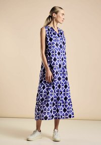 Vestido maxi sin mangas en un patrón geométrico en azul, blanco y negro, con diseño en capas y cierre de botones. Combinado con zapatillas blancas.