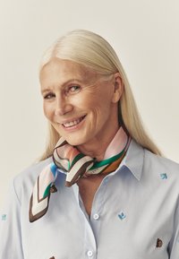 Jedwabny szal z wielokolorowymi wzorami geometrycznymi, w odcieniach różu, zieleni i brązu, noszony na jasnoniebieskiej koszuli z kołnierzykiem, ozdobionej haftem w kwiaty.