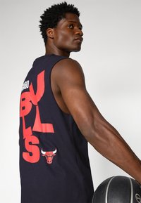 Črna košarkaška majica brez rokavov z velikim rdečim besedilom "CHICAGO BULLS" in majhnim logotipom Bulls na hrbtu. Besedilo je uveljavljeno in grafično.