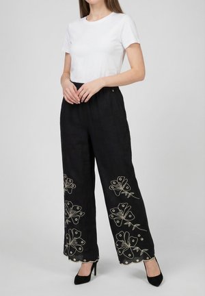 Vrouw draagt zwarte wijde broek met witte bloemenborduursels, gecombineerd met een eenvoudig wit t-shirt met korte mouwen en zwarte hakken.