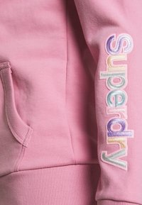 Pink sweatshirt with a soft texture, featuring a side pocket and "Superdry" embroidered in multicolored, iridescent letters on the sleeve.