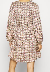 Femme portant une robe de couleur crème avec de longues manches bouffantes et un imprimé floral dense en rose, jaune et noir, vue de dos.