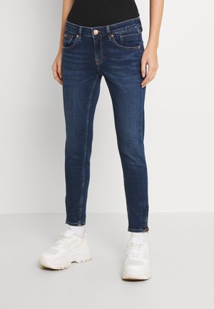 Persona con jeans ajustados de color azul oscuro y zapatillas blancas, de pie frente a un fondo claro y liso.