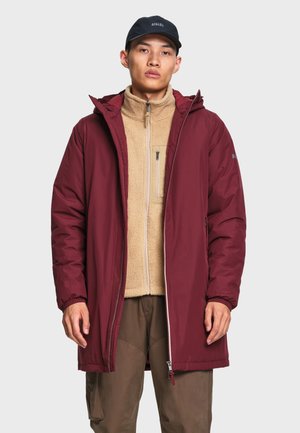 Homme portant un manteau d'hiver à capuche rouge foncé par-dessus une veste polaire beige et un pantalon marron, avec une casquette de baseball bleu foncé sur un fond uni.