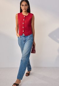 Gilet senza maniche rosso con bottoni dorati, abbinato a jeans a gamba dritta azzurri. L'outfit include sandali con tacco marroni e una borsa bordeaux.
