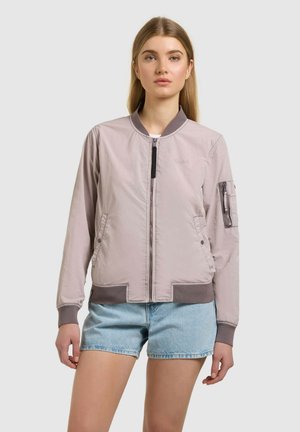 Młoda kobieta w jasnoróżowej kurtce bomberce na zamek błyskawiczny i jasnoniebieskich krótkich spodenkach dżinsowych stoi na tle jednolitego tła.