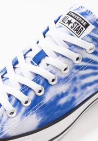 Baskets Converse All Star tie-dye bleu et blanc avec lacets blancs, œillets métalliques et logo de la marque visible sur la languette.