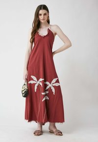 Maxikleid aus burgunderfarbenem Stoff mit ausfransendem Saum. Präsentiert ein Design mit weißen bestickten Palmenblättern. Dünne Träger. Modell hält eine gemusterte Tasche.