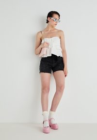 Witte gerimpelde tanktop met een voorknoop, gecombineerd met zwarte denimshorts. Het model draagt roze platform schoenen en witte sokken, en houdt een roze tas vast.