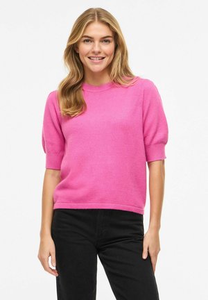 Smiling woman with long blonde hair wearing a bright pink short-sleeve sweater and black pants, standing against a white background.