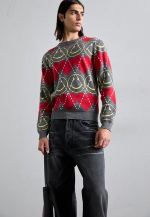 Pull gris avec des motifs géométriques rouges et jaunes, associé à un jeans noir délavé. Il a un col rond et des poignets côtelés.