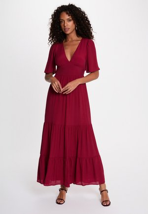Frau mit lockigem Haar, die ein tiefrotes gestuftes Maxikleid mit V-Ausschnitt und schwarze Pumps mit Knöchelriemen trägt, steht vor einem schlichten weißen Hintergrund.