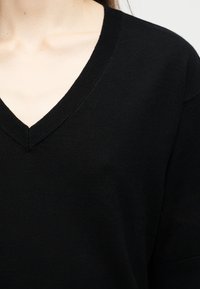 Κοντινό πλάνο ενός ατόμου που φοράει μαύρο πουλόβερ με V λαιμόκοψη, εμφανίζοντας το λαιμό και το πάνω μέρος του στήθους.