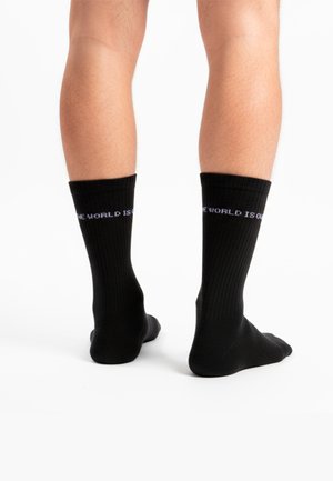 Crne pamučne čarape s rebrastom teksturom. Sadrže bijeli natpis 'SVIJET JE NAŠ' na rubu. Vidljive su na nogama osobe.