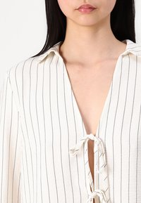 Femme portant un chemisier blanc à fines rayures verticales noires, avec un col et des nœuds noués le long d'une ouverture en V.