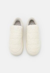 Dvojice světle béžových chlupatých slip-on tenisek s gumovými podrážkami, pohled shora na jednobarevném bílém pozadí.