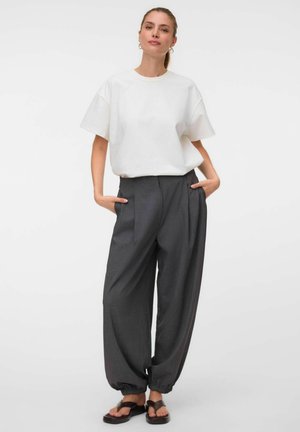 Femme portant une chemise blanche ample à manches courtes, un pantalon large gris foncé avec des poignets élastiques, et des sandales noires ouvertes aux orteils, debout les mains dans les poches.