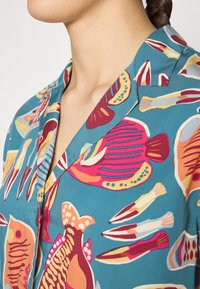 Camicia a maniche corte con sfondo turchese e motivi di pesci e pennelli in colori vivaci, texture liscia e colletto a bavero.