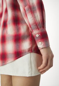 Gros plan d'une personne portant une chemise à manches longues à carreaux rouges avec des poignets boutonnés, associée à une jupe blanche, montrant le côté et la main.