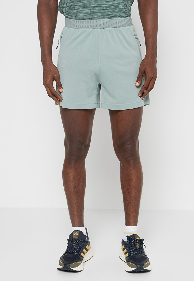 Pantalones cortos deportivos de color verde claro con cintura elástica, bolsillos laterales y una textura suave, combinados con zapatillas marinas con acentos amarillos.