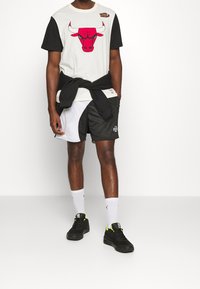 Bílý tričko s grafickým motivem červeného býka a černými rukávy, spárované s černobílými šortkami. Dokončeno s černými teniskami a bílými ponožkami.