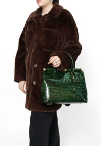 Borsa verde in ecopelle a motivo coccodrillo con un design strutturato, dettagli in hardware dorato e manico corto. Indossata con un cappotto oversized in pelliccia sintetica marrone.