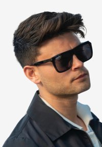 Svarta rektangulära solglasögon med tjocka bågar, bärs av en person. De har en glansig yta och tonade linser. Bakgrunden är helt vit.