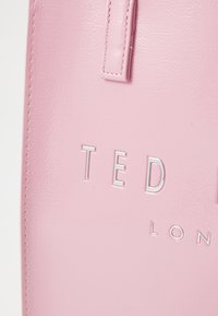 Vaaleanpunainen tekonahkainen laukku, jossa on kohokuvioitu hopeinen teksti "TED" ja "LONDON." Sujuva pinta kahdella yläkahvalla.