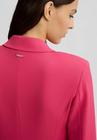 Blazer fuchsia avec un col cranté et une texture lisse. Doté d'un petit accent argenté au niveau de l'encolure arrière. Coupe ajustée.