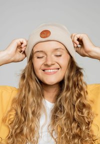 Beżowa wełniana czapka z brązową skórzaną naszywką z wzorem; modelka z kręconymi włosami w żółtej kurtce i białej koszuli.