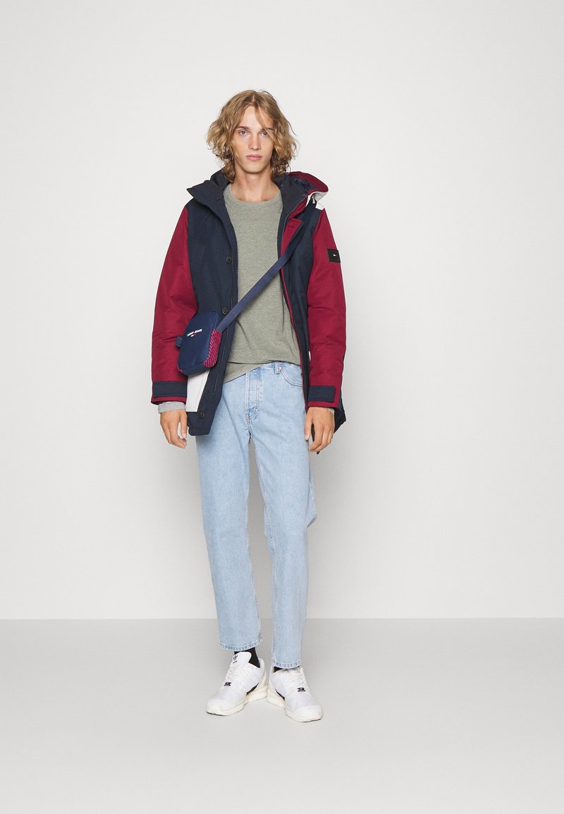 Marineblå og burgunder fargeblockert vanntett jakke med hette, grå t-skjorte, lys blå jeans, og hvite sneakers; crossbody-veske inkludert.